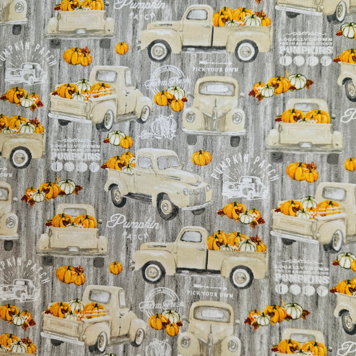 A repeating fabric or wallpaper pattern featuring vintage cream pickup trucks carrying orange pumpkins and gourds on a weathered gray background with faint white decorative text.