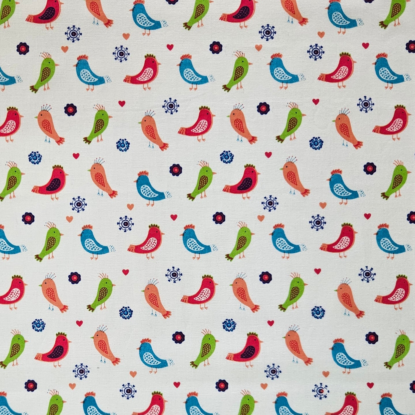 A close-up of a fabric pattern featuring colourful stylized birds in red, blue, green, and orange on a warm white background, accented with small red hearts and dark blue floral motifs.