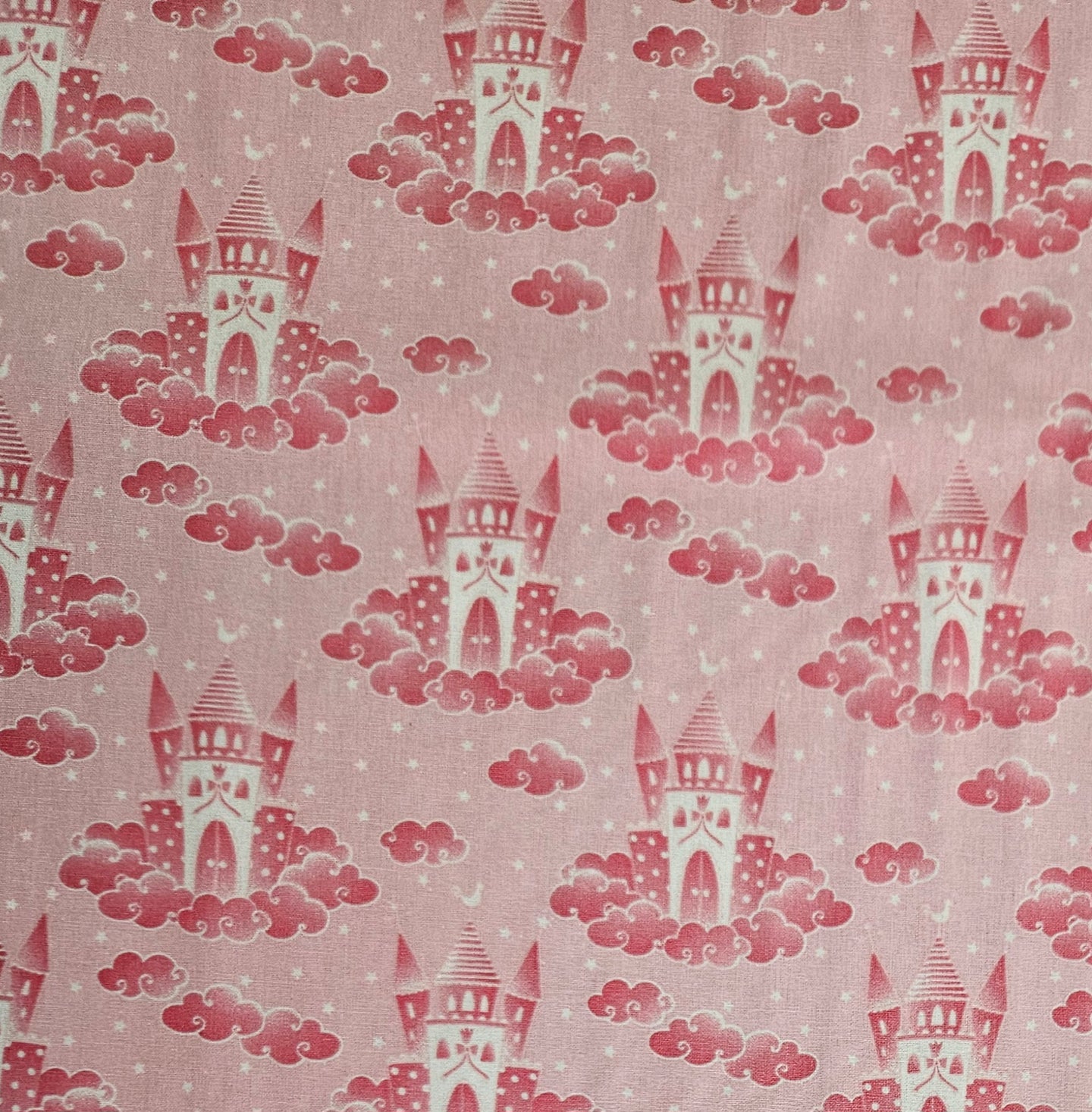 A close-up of a pink fabric with a repeating pattern of red castles rising from pink clouds, with small white stars scattered throughout.