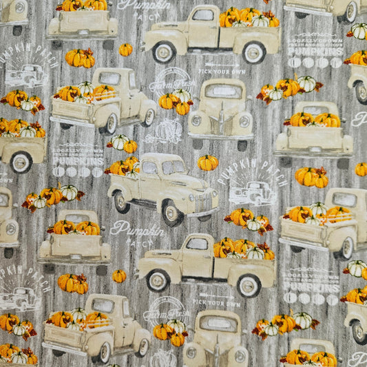 A repeating fabric or wallpaper pattern featuring vintage cream pickup trucks carrying orange pumpkins and gourds on a weathered gray background with faint white decorative text.